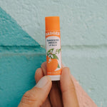 Badger Tangerine Breeze Lip Balm held against a colourful wall