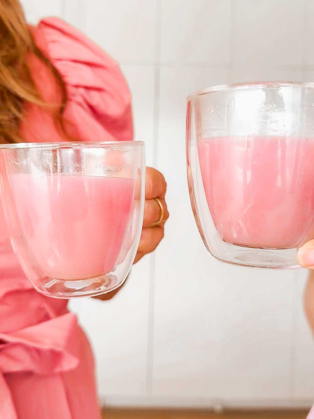 Two people clink double-walled glass cups filled with Supercharged Food Natural Silica The Beauty Mineral Vegan Collagen Support. One wears a pink puff-sleeve dress against a light, minimal background.