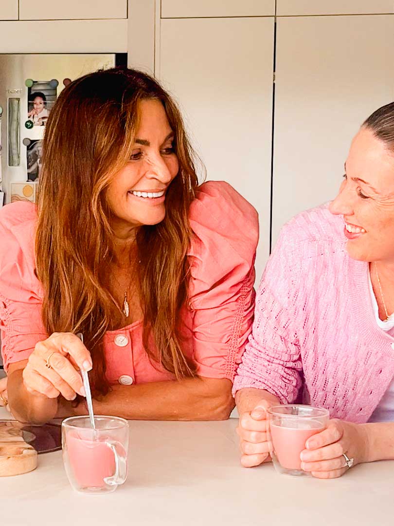 Two women in pink tops smile and chat at a kitchen counter, each holding a glass of Supercharged Food Natural Silica The Beauty Mineral Vegan Collagen Support, enjoying the collagen-boosting supplement together.