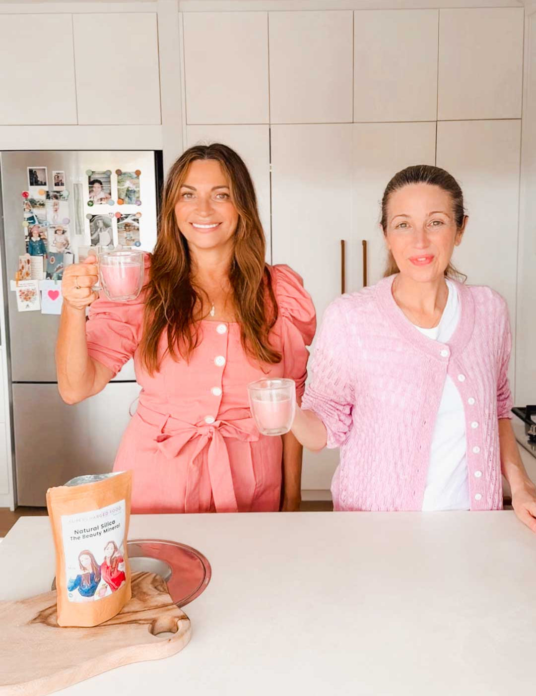 Two women in pink outfits smile and hold mugs in a bright kitchen, with a package of Supercharged Food Natural Silica The Beauty Mineral Vegan Collagen Support 90g on the counter. The fridge behind them is decorated with family photos and magnets.