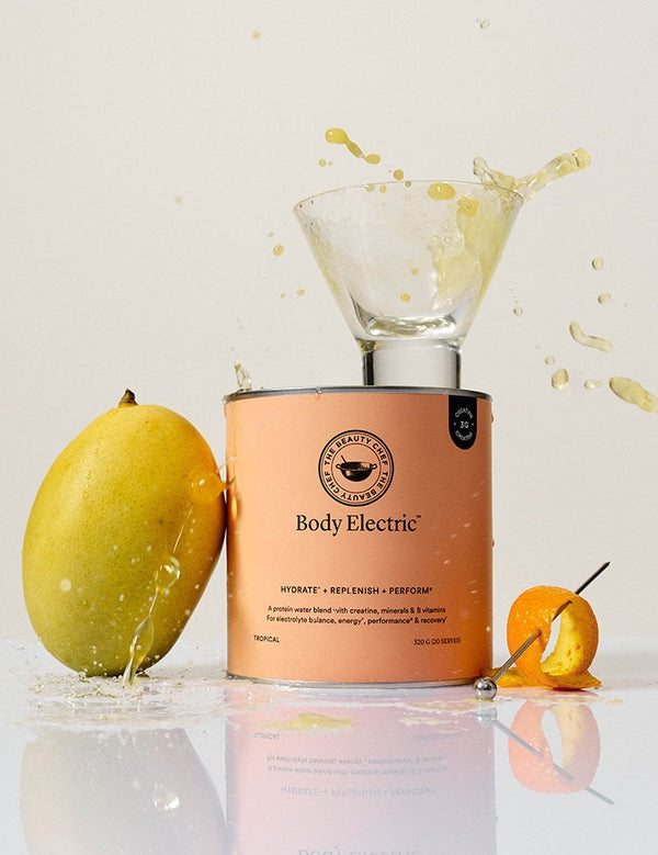 The Beauty Chef Body Electric Tropical canister beside a whole mango and curled orange peel, with a small glass on top and pale-yellow liquid splashing up from the glass.
