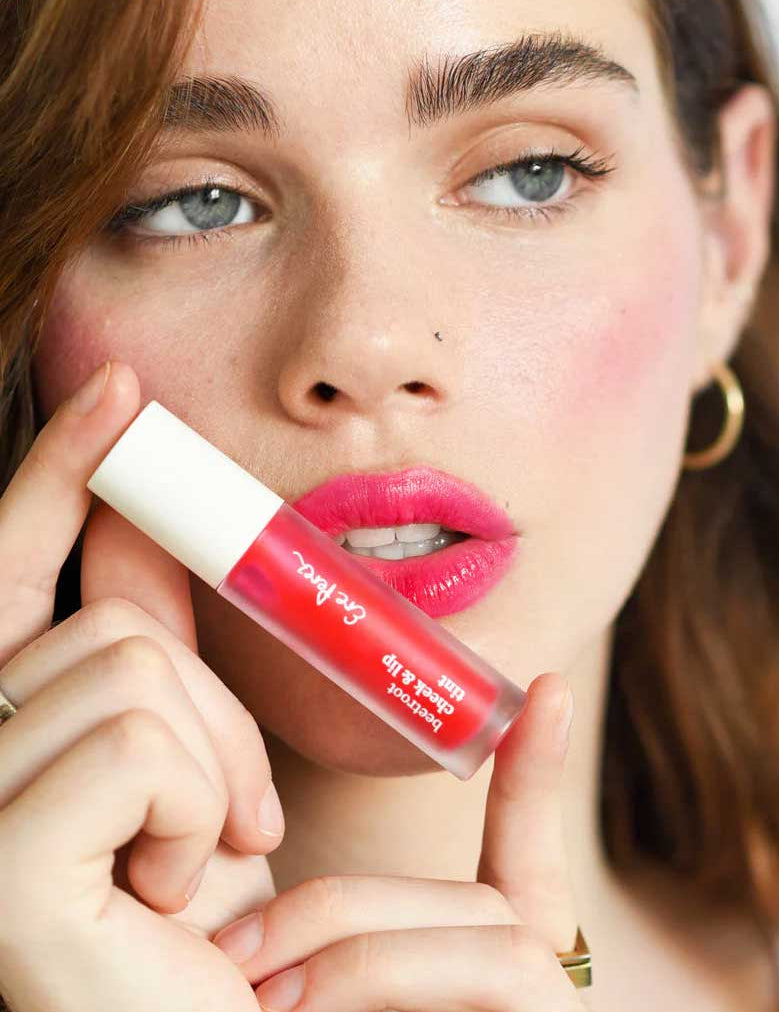 A fair-skinned woman with brown hair holds Ere Perez Beetroot Cheek & Lip Tint 4.5ml near her lips, wearing bright pink lipstick, gold hoop earrings, and a gold ring against a soft-focus background.