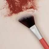 A makeup brush with a red handle lies next to a pile of loose, crushed Benecos Beauty ID Palette Blush Refills on a light surface.