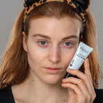 Model holding a tube of Benecos Face Primer beside her cheek while wearing a playful leopard-print cat-ear headband.