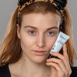 Model holding a tube of Benecos Face Primer beside her cheek while wearing a playful leopard-print cat-ear headband.