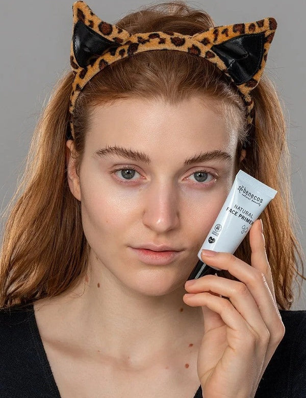 Model holding a tube of Benecos Face Primer beside her cheek while wearing a playful leopard-print cat-ear headband.