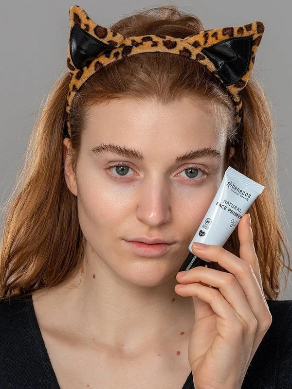 Model holding a tube of Benecos Face Primer beside her cheek while wearing a playful leopard-print cat-ear headband.