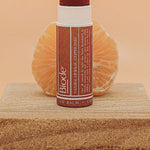 Uncapped Biode Copper Daisy balm in a brown paper tube on a stone block, exposing the copper-coloured stick. A peeled mandarin sits behind it, with Copper Daisy printed below.