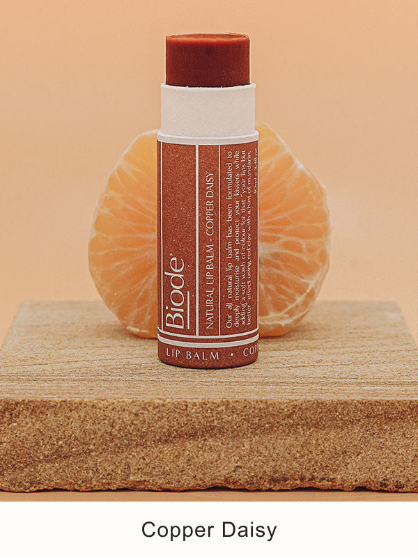 Uncapped Biode Copper Daisy balm in a brown paper tube on a stone block, exposing the copper-coloured stick. A peeled mandarin sits behind it, with Copper Daisy printed below.
