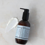 Biode Cream Cleanser with Rosemary 200ml amber pump bottle beside a smear of white cream cleanser on a pale stone surface.