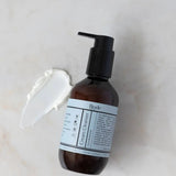 Biode Cream Cleanser with Rosemary 200ml amber pump bottle beside a smear of white cream cleanser on a pale stone surface.