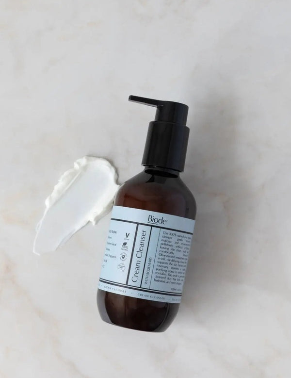 Biode Cream Cleanser with Rosemary 200ml amber pump bottle beside a smear of white cream cleanser on a pale stone surface.