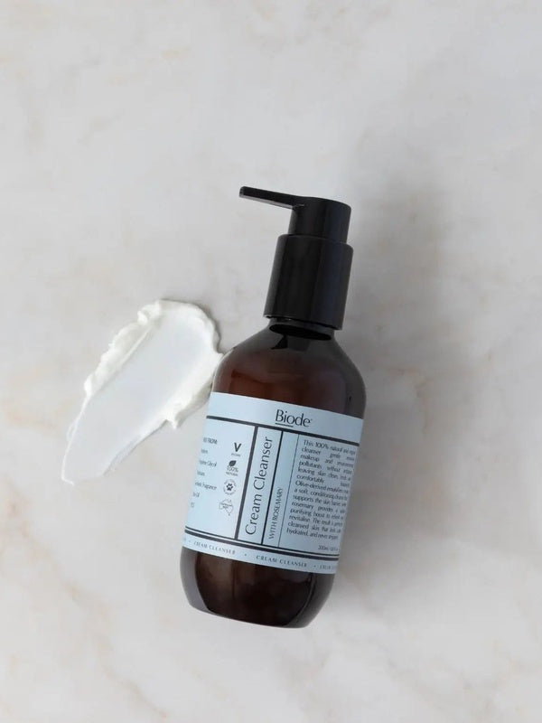 Biode Cream Cleanser with Rosemary 200ml amber pump bottle beside a smear of white cream cleanser on a pale stone surface.