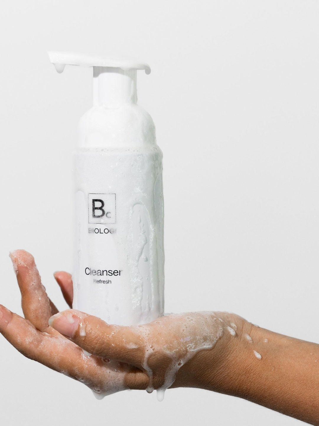 A hand supports a Biologi Bc Refresh Cleanser pump bottle covered with foam and water droplets, against a pale studio background.