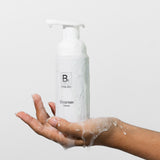 A hand supports a Biologi Bc Refresh Cleanser pump bottle covered with foam and water droplets, against a pale studio background.