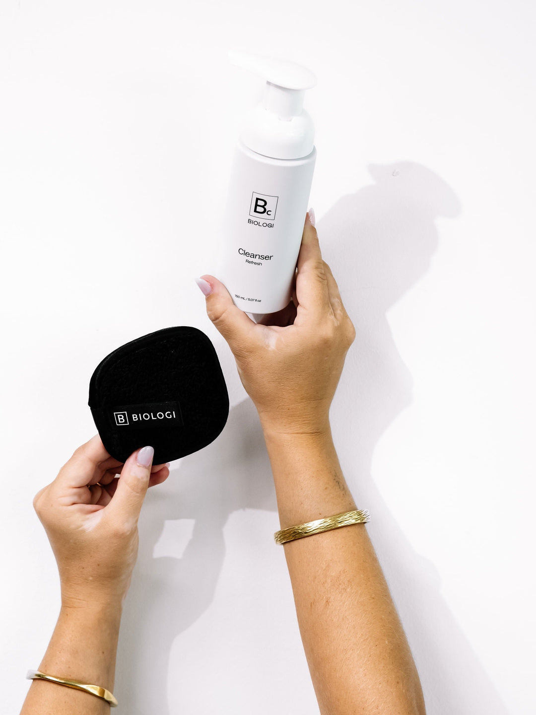 Hands hold Biologi Bc Refresh Cleanser and a round black Biologi cleansing mitt against white, showing a cleansing routine with the accessory.