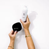Hands hold Biologi Bc Refresh Cleanser and a round black Biologi cleansing mitt against white, showing a cleansing routine with the accessory.
