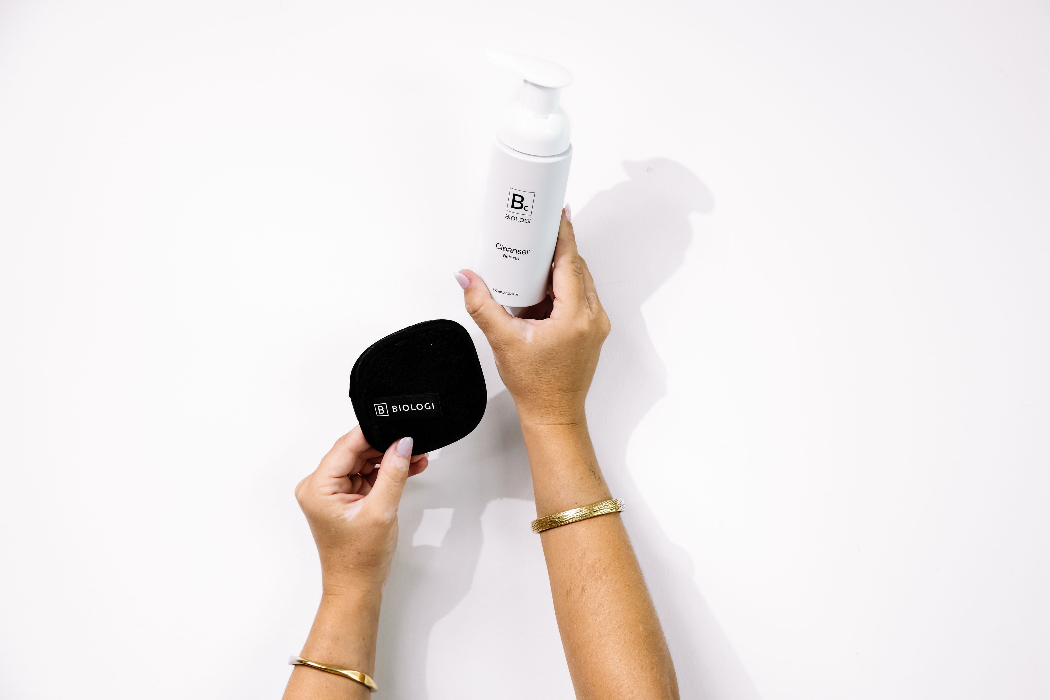 Hands hold Biologi Bc Refresh Cleanser and a round black Biologi cleansing mitt against white, showing a cleansing routine with the accessory.