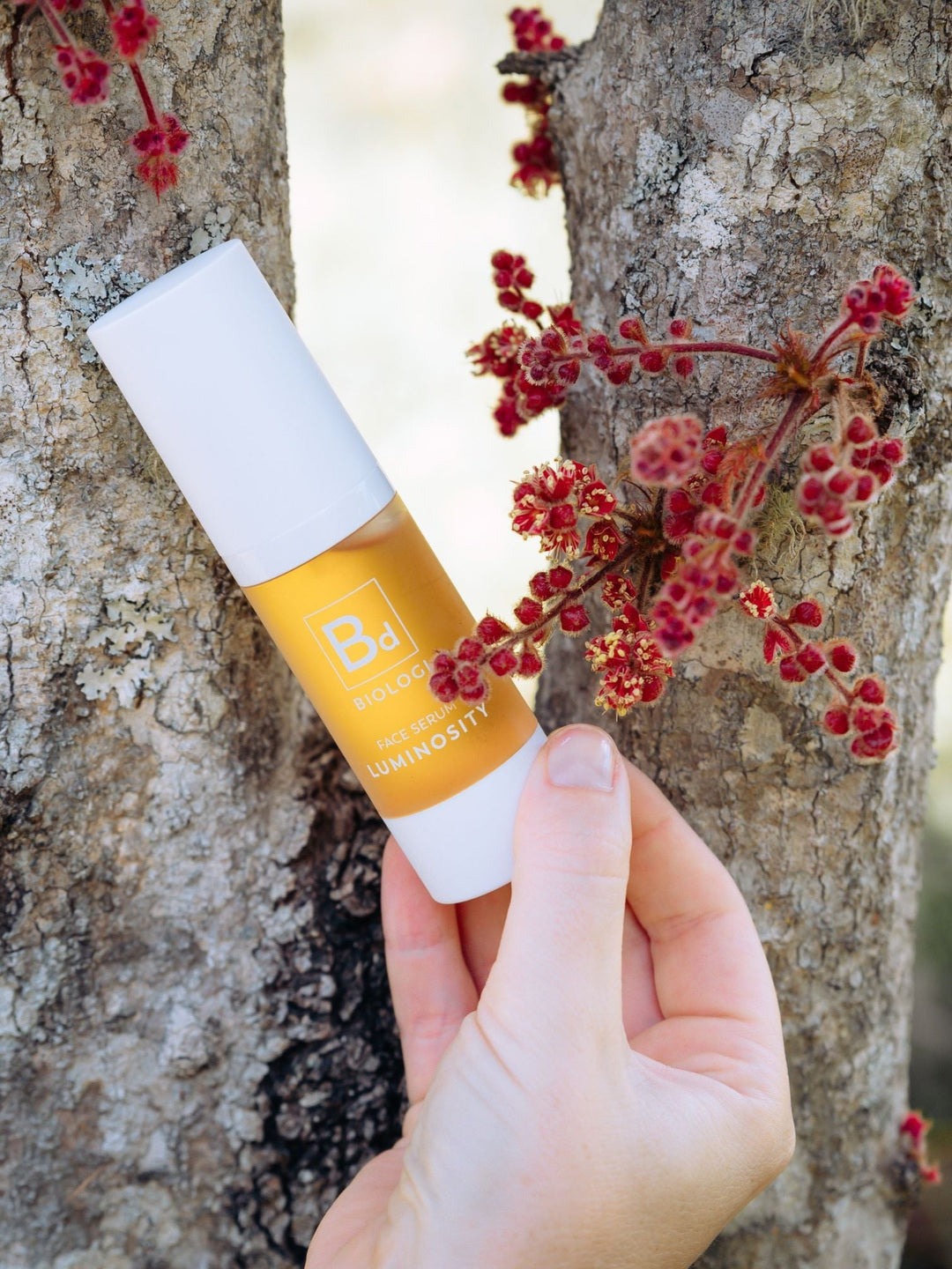 A hand holds a white-capped Biologi Bd Luminosity Face Serum bottle beside a tree trunk and small red botanical clusters outdoors.