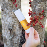 A hand holds a white-capped Biologi Bd Luminosity Face Serum bottle beside a tree trunk and small red botanical clusters outdoors.