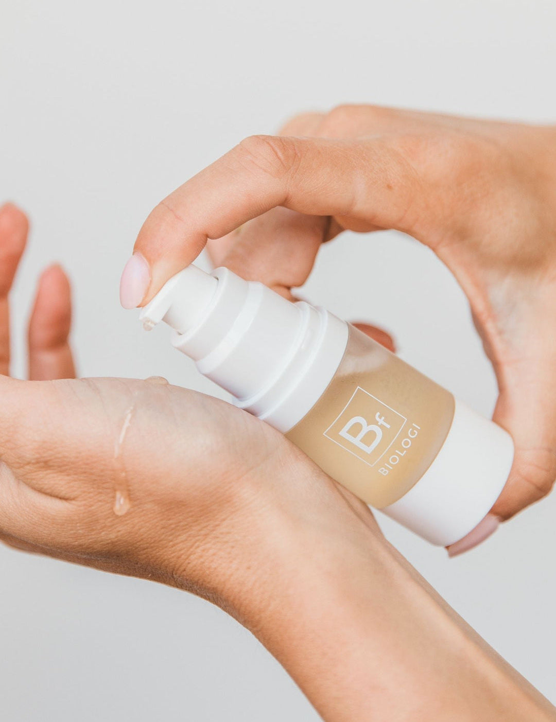 Hands dispensing Biologi Bf Restore Face & Body Serum from the white pump onto the back of a hand, showing the golden serum and how it is applied.