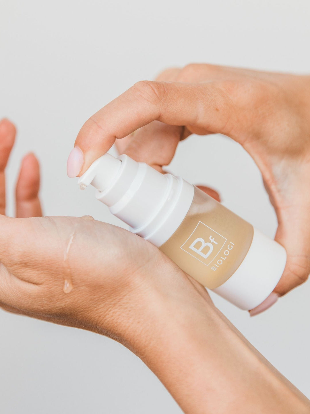 Hands dispensing Biologi Bf Restore Face & Body Serum from the white pump onto the back of a hand, showing the golden serum and how it is applied.