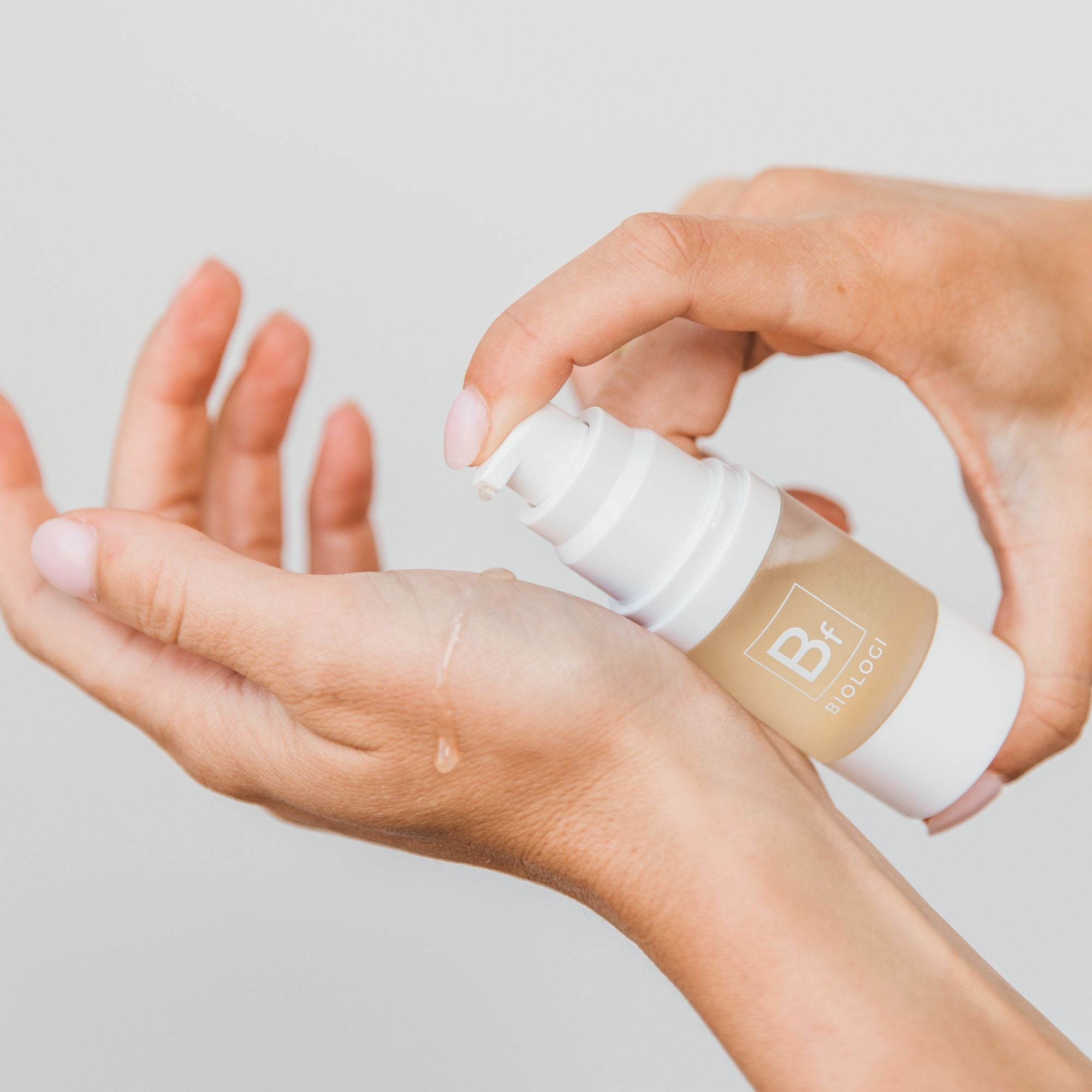 Hands dispensing Biologi Bf Restore Face & Body Serum from the white pump onto the back of a hand, showing the golden serum and how it is applied.