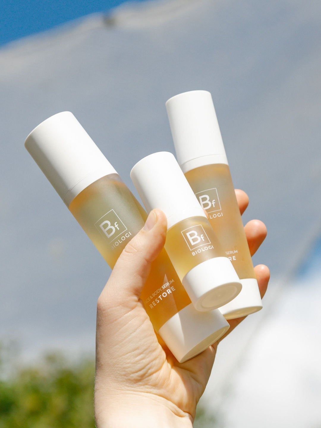 A hand holds three sizes of Biologi Bf Restore Face & Body Serum outdoors against blue sky and a white fabric backdrop.