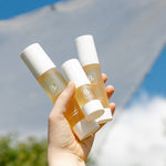 A hand holds three sizes of Biologi Bf Restore Face & Body Serum outdoors against blue sky and a white fabric backdrop.