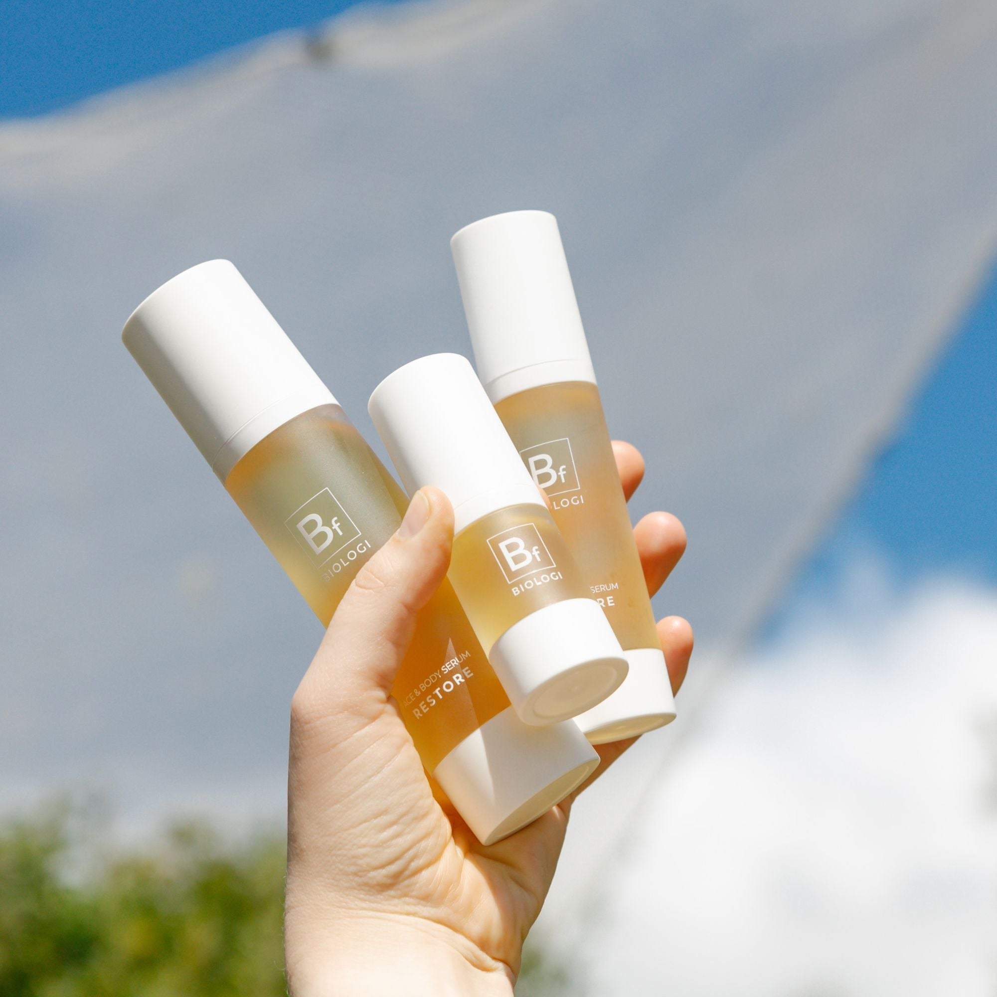 A hand holds three sizes of Biologi Bf Restore Face & Body Serum outdoors against blue sky and a white fabric backdrop.