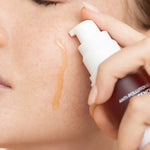 Close-up of Biologi Bg Defence Anti-Pollution Serum being dispensed beside a woman's cheek, with a drop of amber serum running down the skin.