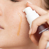Close-up of Biologi Bg Defence Anti-Pollution Serum being dispensed beside a woman's cheek, with a drop of amber serum running down the skin.