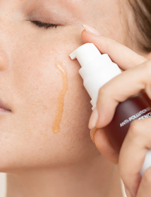 Close-up of Biologi Bg Defence Anti-Pollution Serum being dispensed beside a woman's cheek, with a drop of amber serum running down the skin.