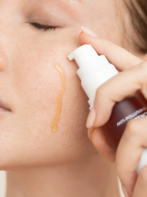 Close-up of Biologi Bg Defence Anti-Pollution Serum being dispensed beside a woman's cheek, with a drop of amber serum running down the skin.