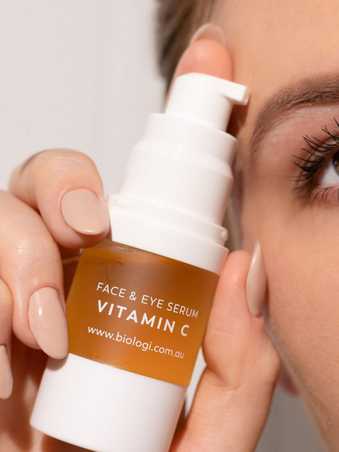 A woman holds an uncapped Biologi Bk Vitamin C Face & Eye Serum pump bottle beside her eye, with the label and white dispensing pump visible.