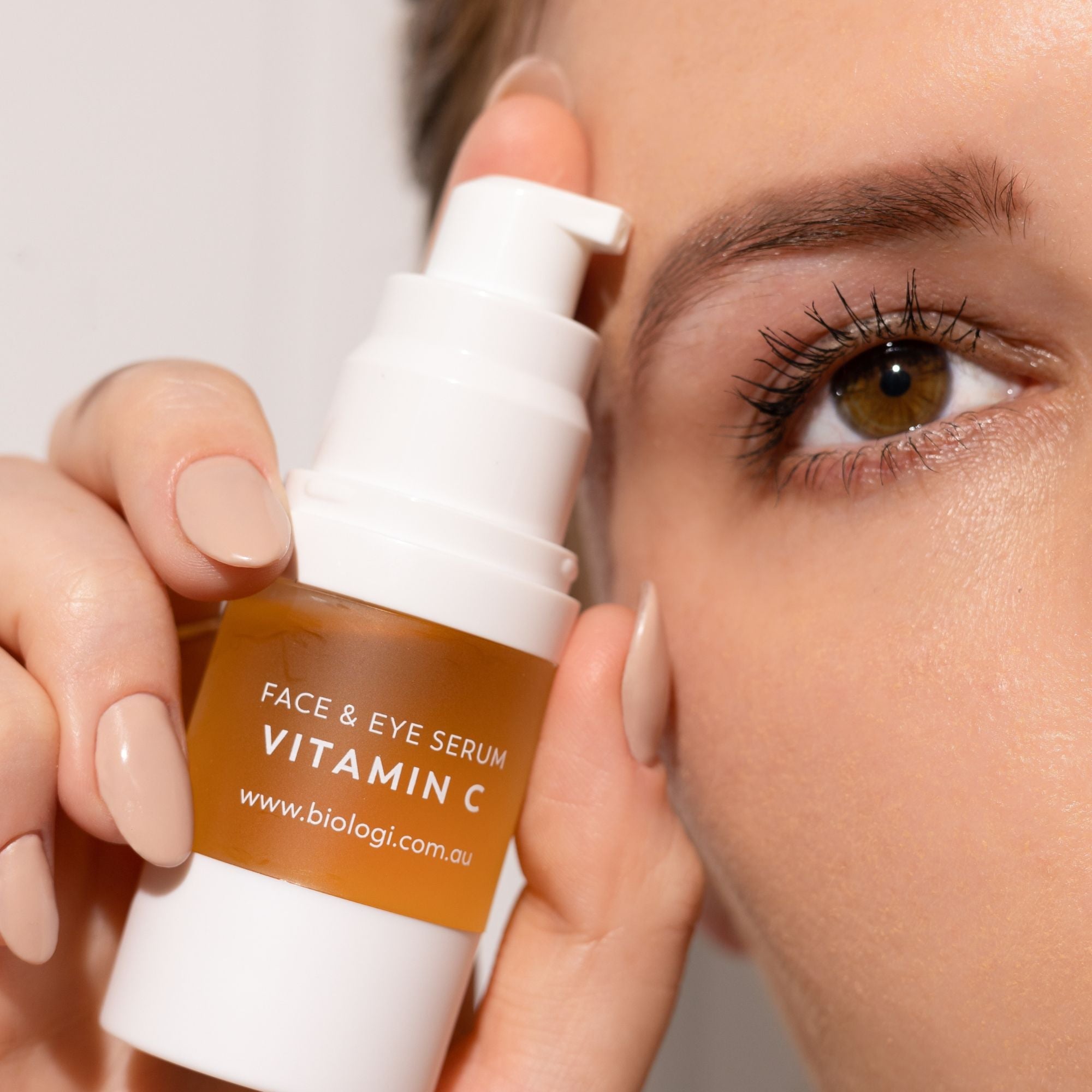 A woman holds an uncapped Biologi Bk Vitamin C Face & Eye Serum pump bottle beside her eye, with the label and white dispensing pump visible.