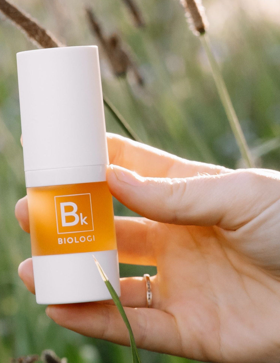 A hand displays the white-capped Biologi Bk Vitamin C Face & Eye Serum bottle in sunlight among soft green foliage.