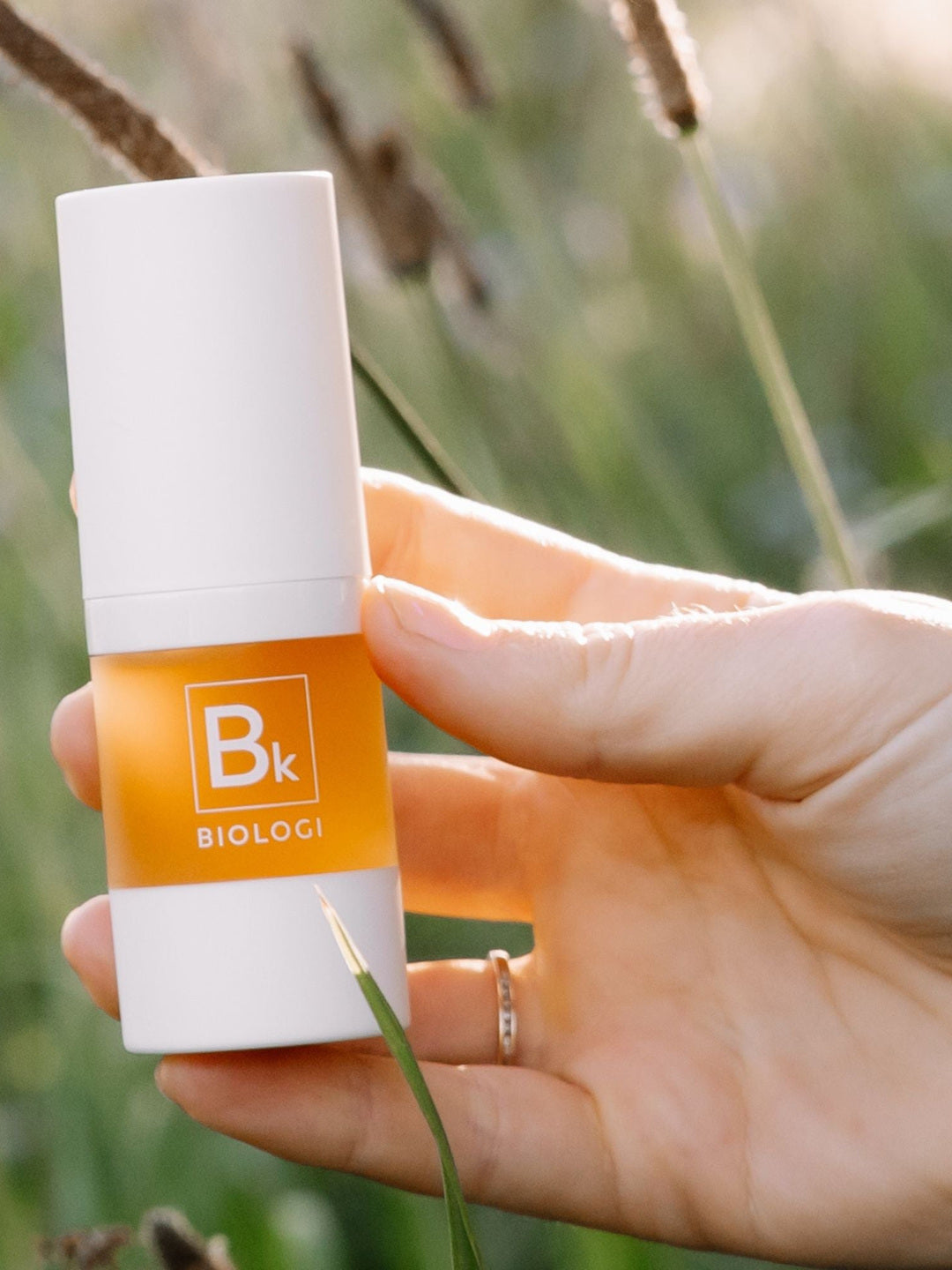 A hand displays the white-capped Biologi Bk Vitamin C Face & Eye Serum bottle in sunlight among soft green foliage.
