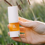 A hand displays the white-capped Biologi Bk Vitamin C Face & Eye Serum bottle in sunlight among soft green foliage.
