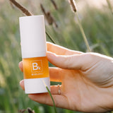 A hand displays the white-capped Biologi Bk Vitamin C Face & Eye Serum bottle in sunlight among soft green foliage.