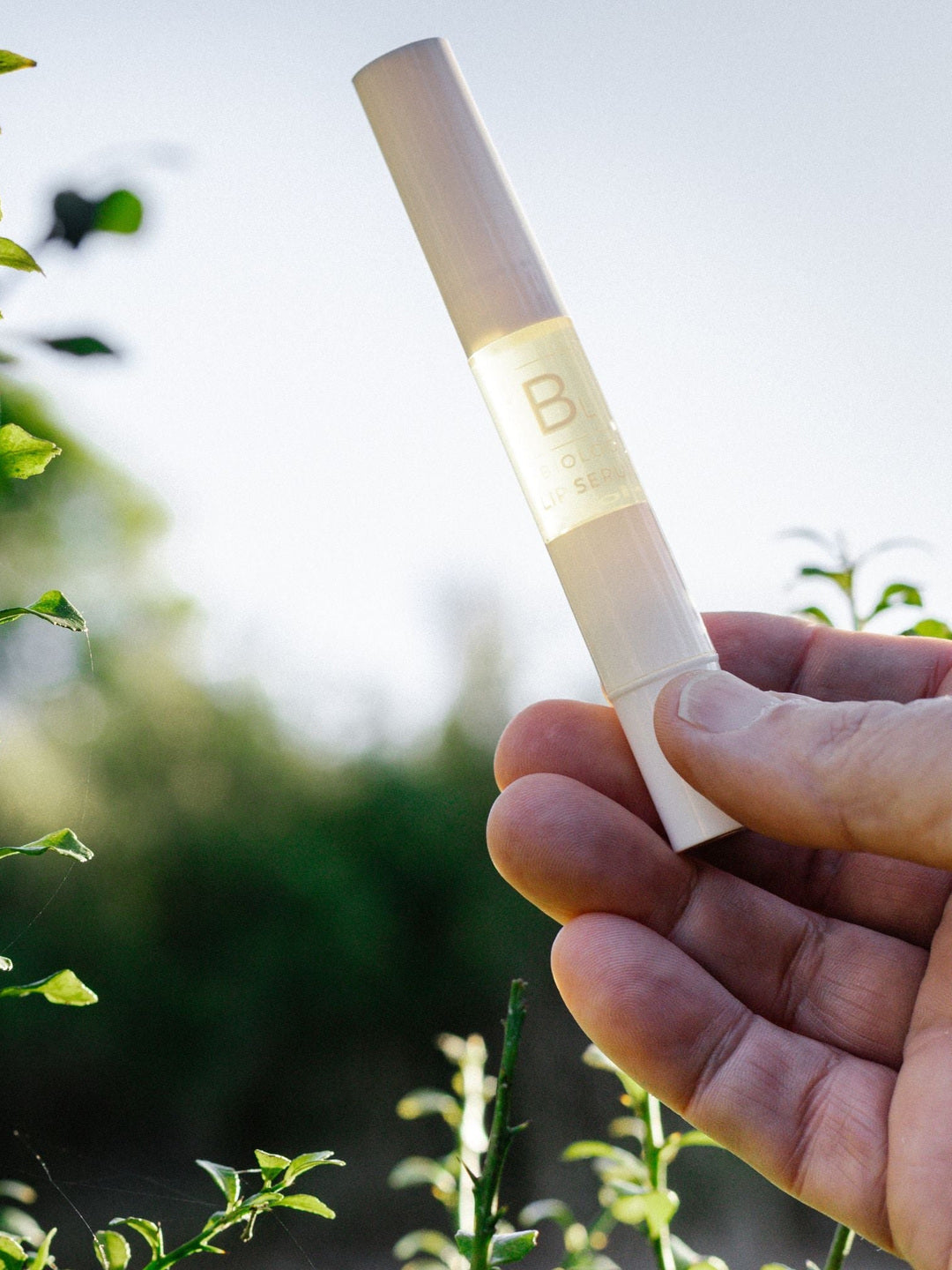 A hand holds the slim capped Biologi Bl Nourish Lip Serum pen against blue sky and green plants in bright sunlight.