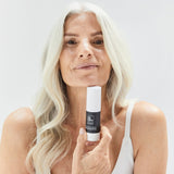 A woman with long grey-blonde hair holds Biologi Bm Regenerate Anti-Ageing Serum beneath her chin, with the white bottle and deep plum label facing forward.