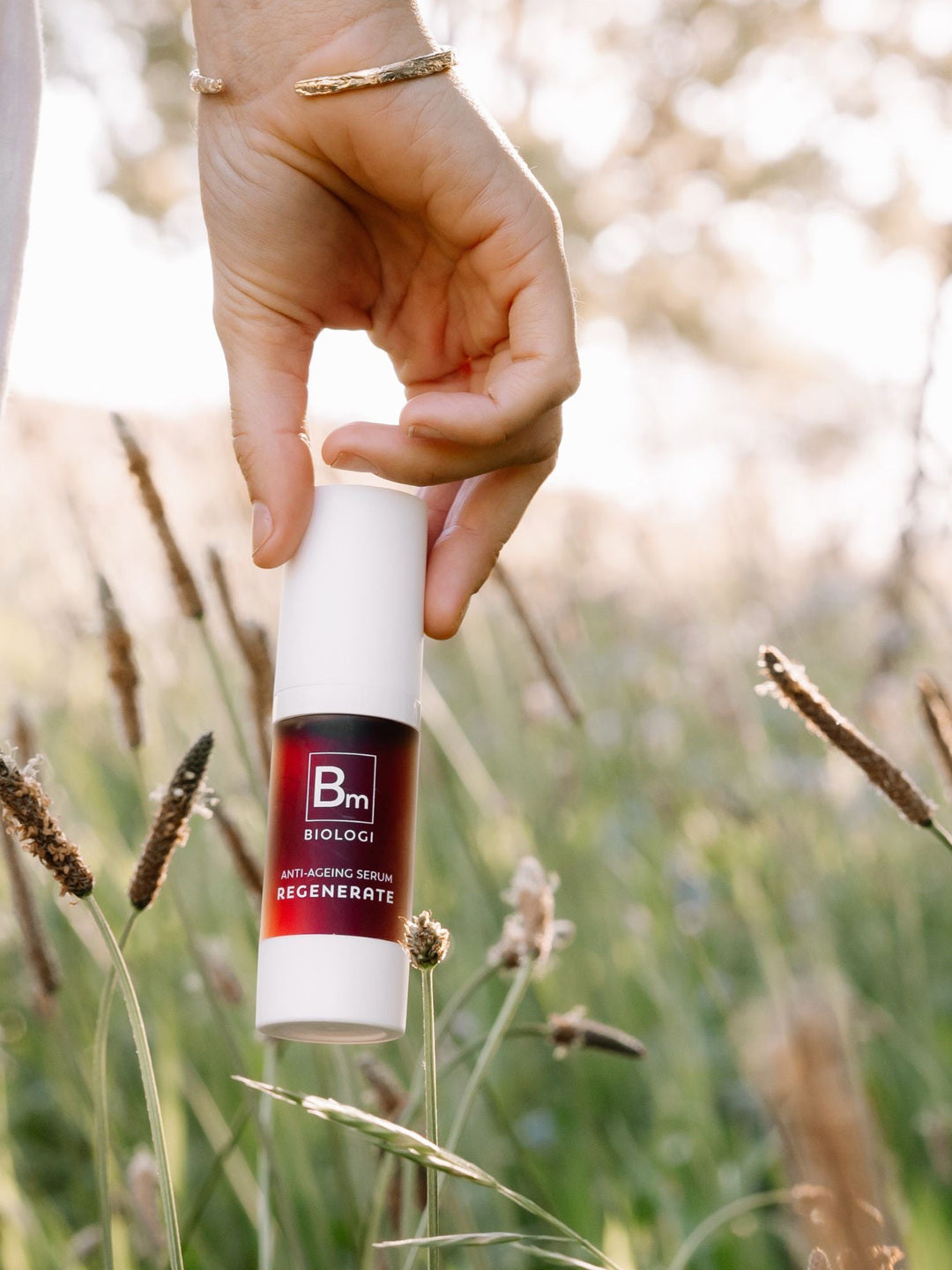 A hand holds the capped Biologi Bm Regenerate Anti-Ageing Serum bottle among sunlit grasses, showing its white packaging and dark plum label.