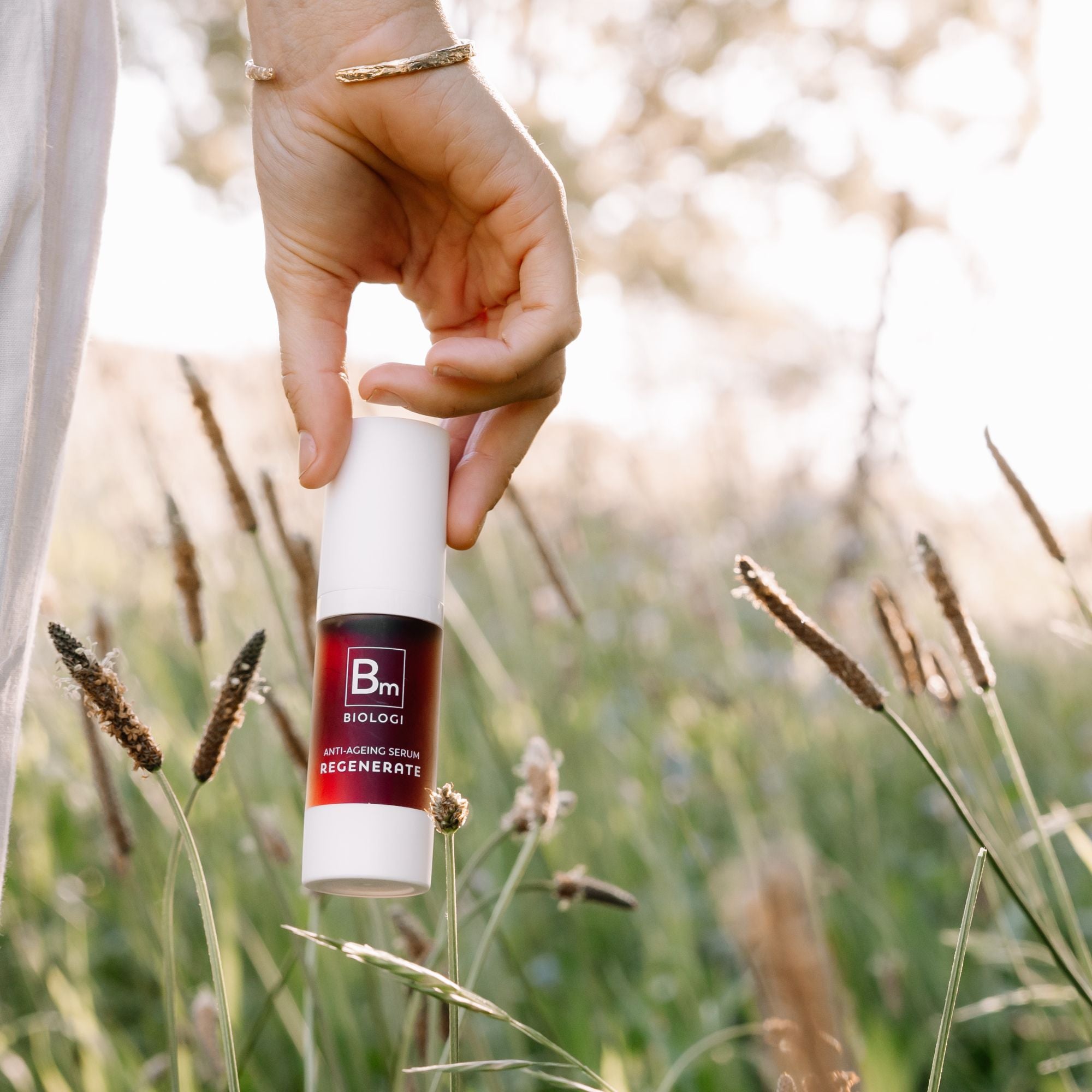 A hand holds the capped Biologi Bm Regenerate Anti-Ageing Serum bottle among sunlit grasses, showing its white packaging and dark plum label.