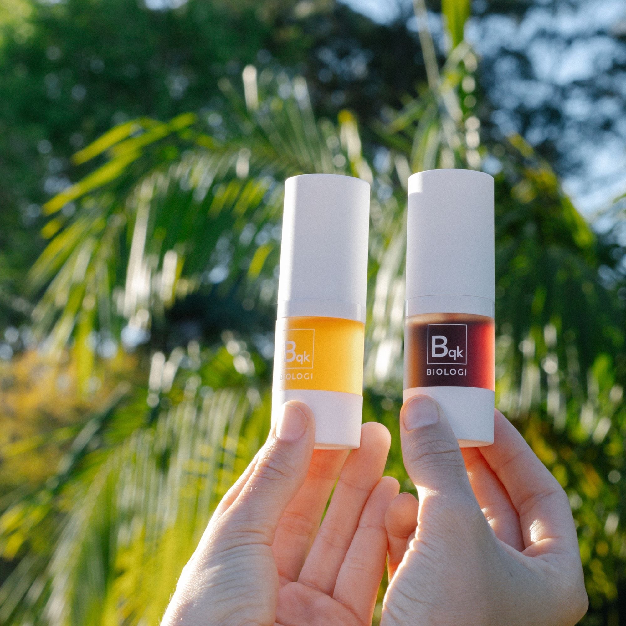 Two hands hold the Biologi Bqk Radiance Face Serum Duo outdoors against sunlit palm leaves, showing both capped serum bottles side by side.