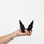 A hand holds Biologi Br Rosehip Oil and By Blackberry Seed Oil together, showing the two black dropper bottles against a pale background.