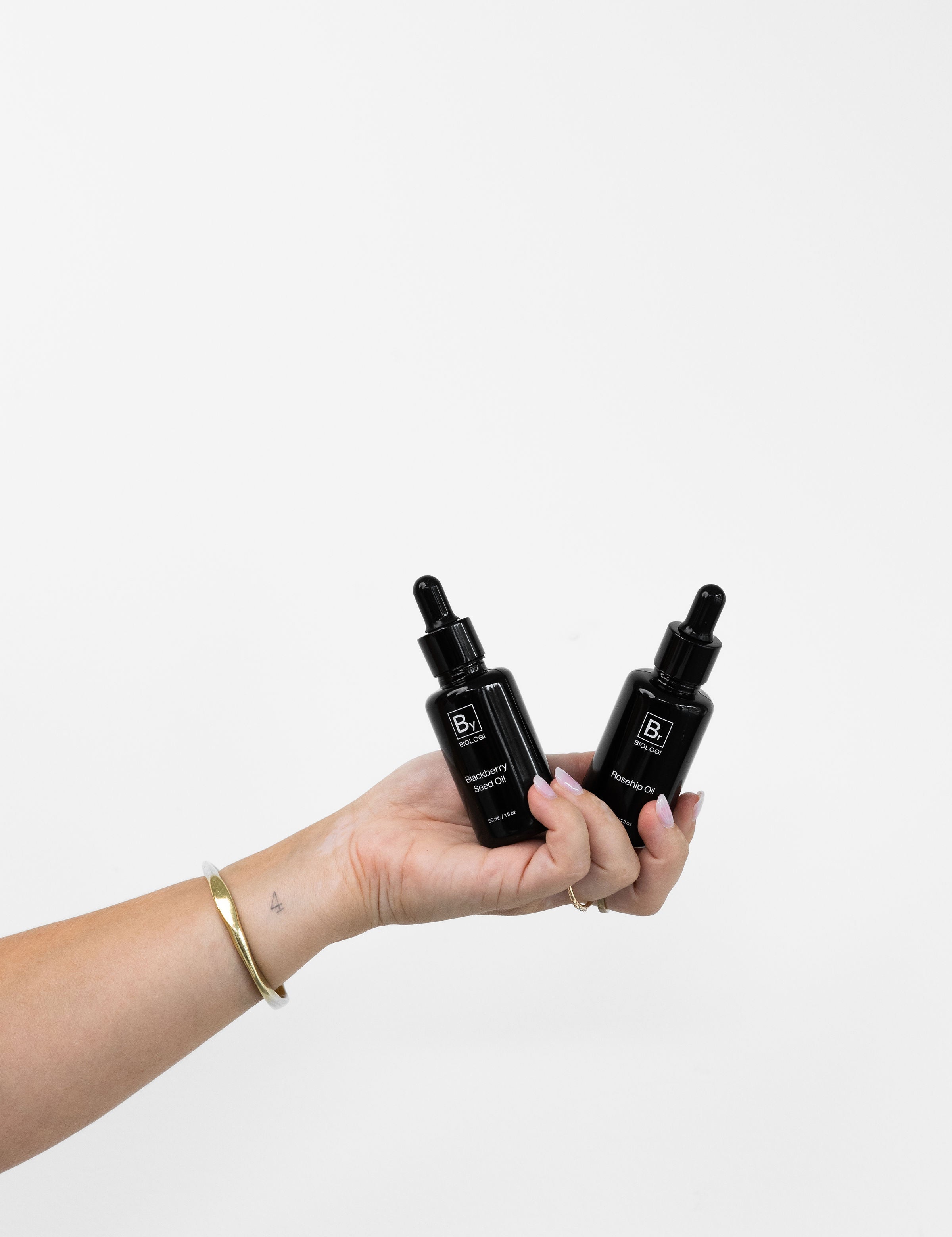 A hand holds Biologi Br Rosehip Oil and By Blackberry Seed Oil together, showing the two black dropper bottles against a pale background.