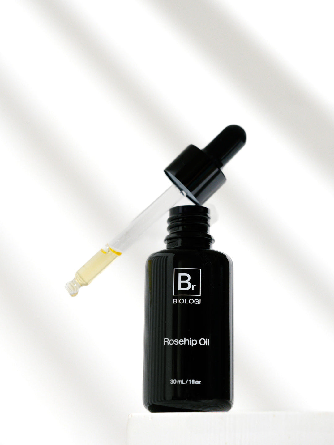 An open Biologi Br Rosehip Oil bottle stands on a white block beneath its raised glass dropper containing pale yellow oil, with soft diagonal shadows.