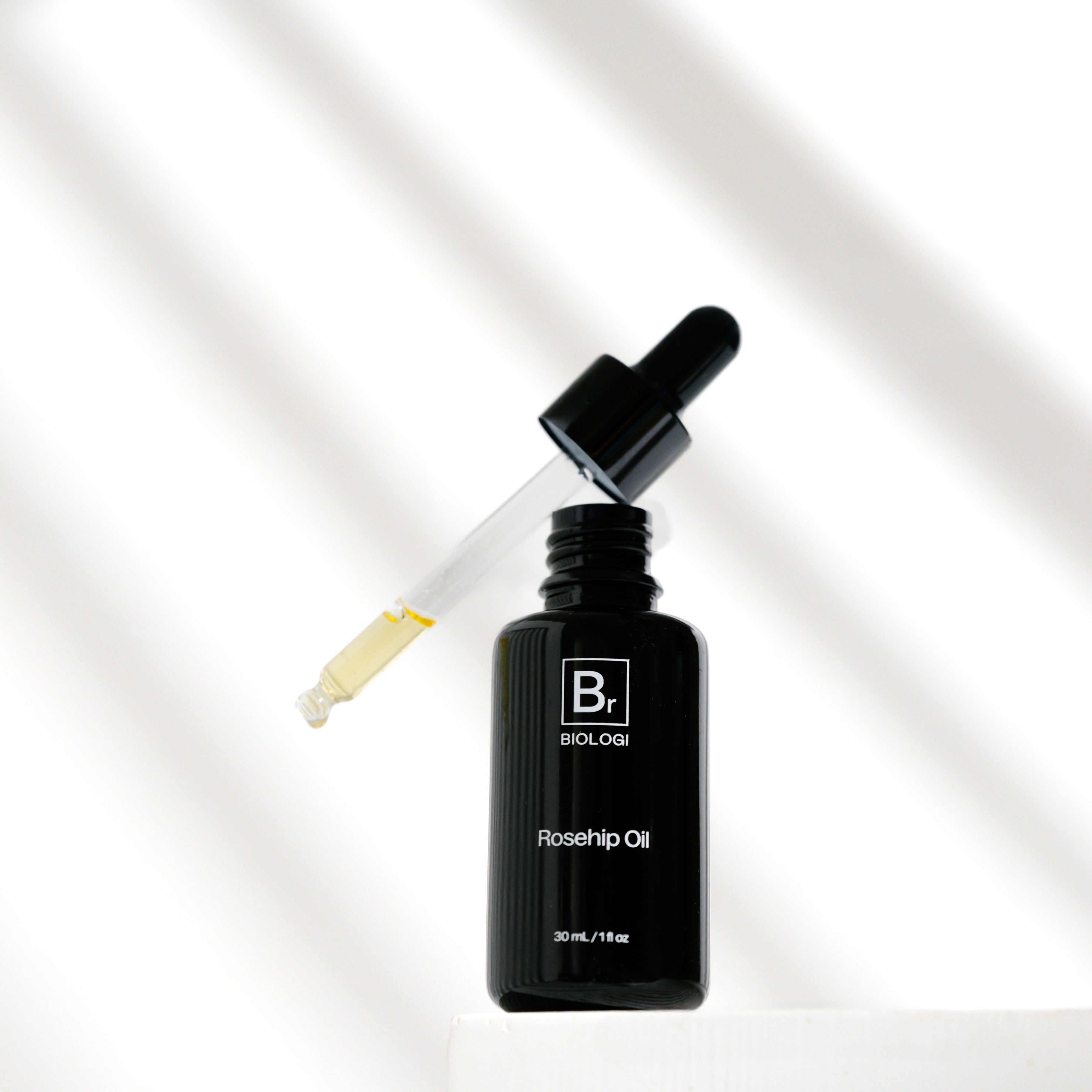 An open Biologi Br Rosehip Oil bottle stands on a white block beneath its raised glass dropper containing pale yellow oil, with soft diagonal shadows.