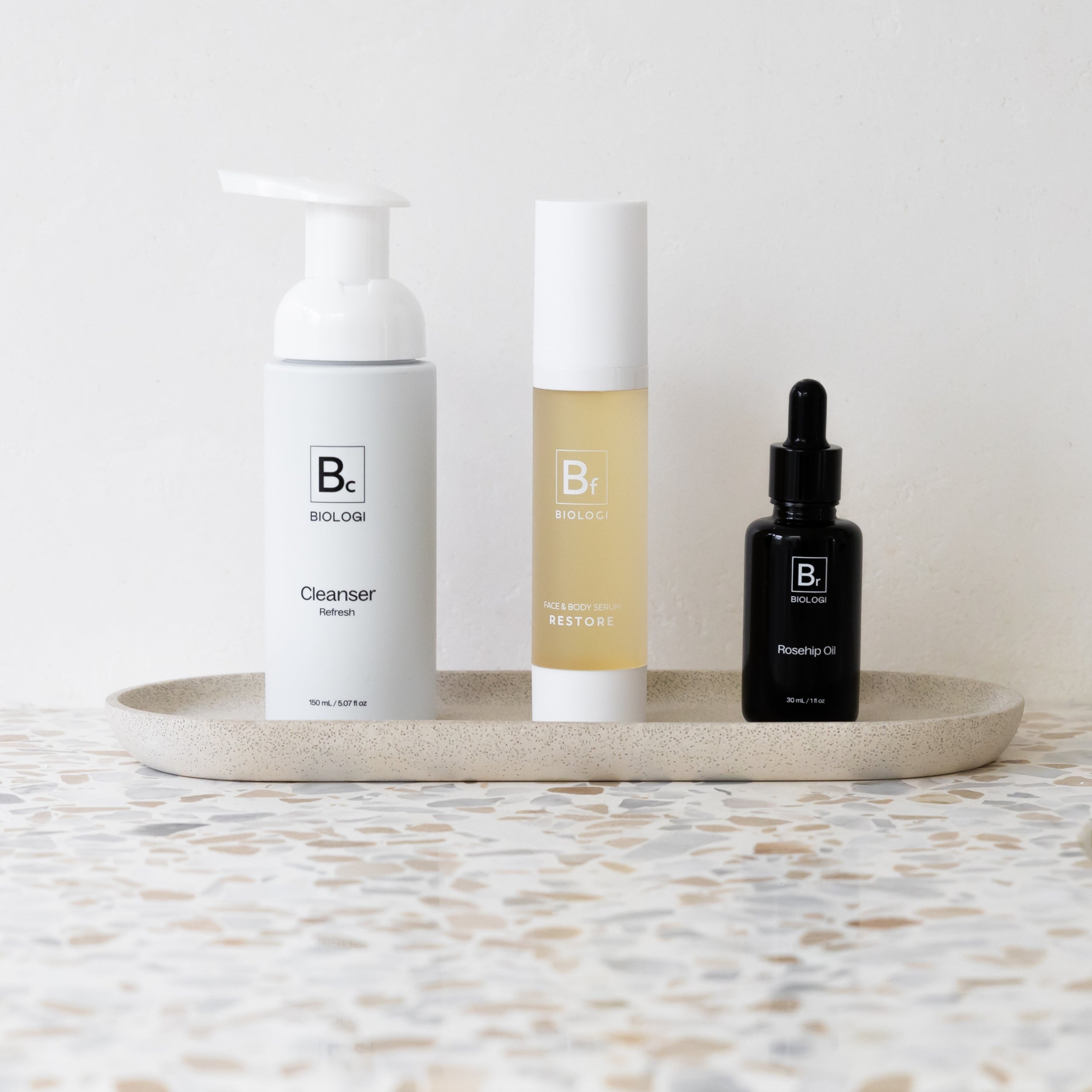Biologi Bc Refresh Cleanser, Bf Restore serum and Br Rosehip Oil arranged together on a stone tray, showing the three products in a skincare routine.